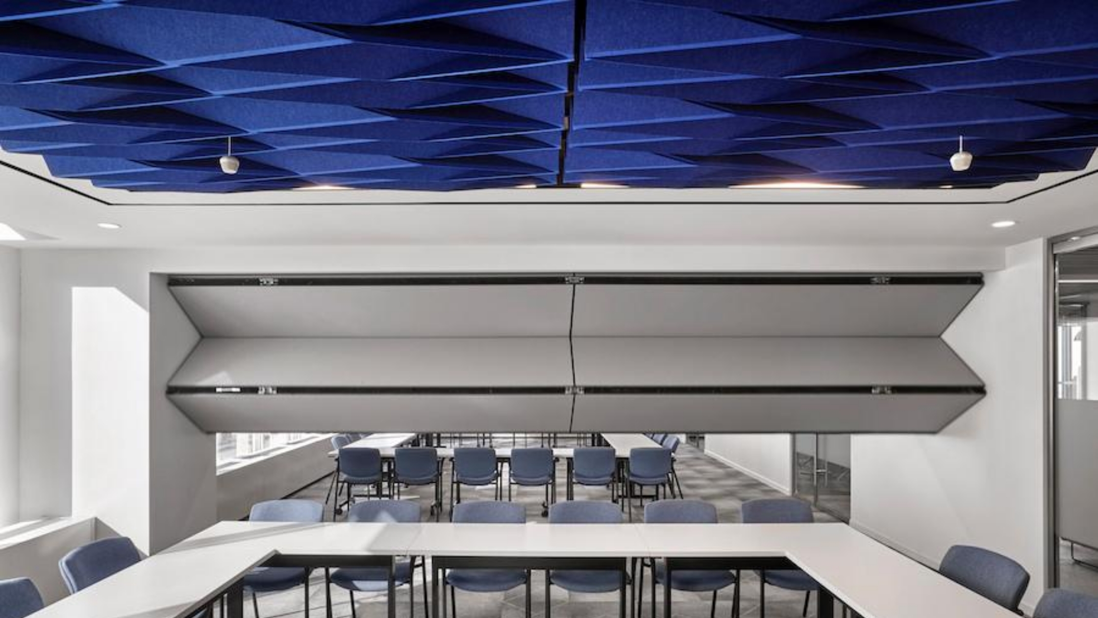 White Skyfold Classic operable partition folding from the ceiling to subdivide an office conference room that has blue acoustic ceiling baffles.